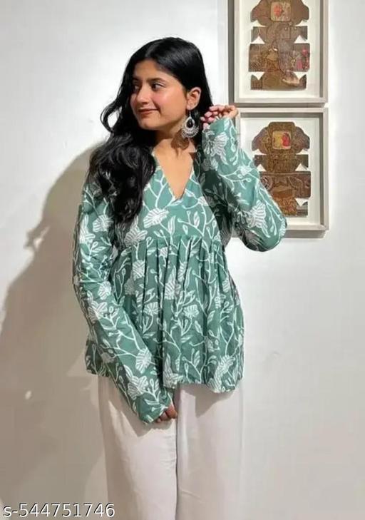 Woman wearing a green floral blouse with white pants against a white wall.